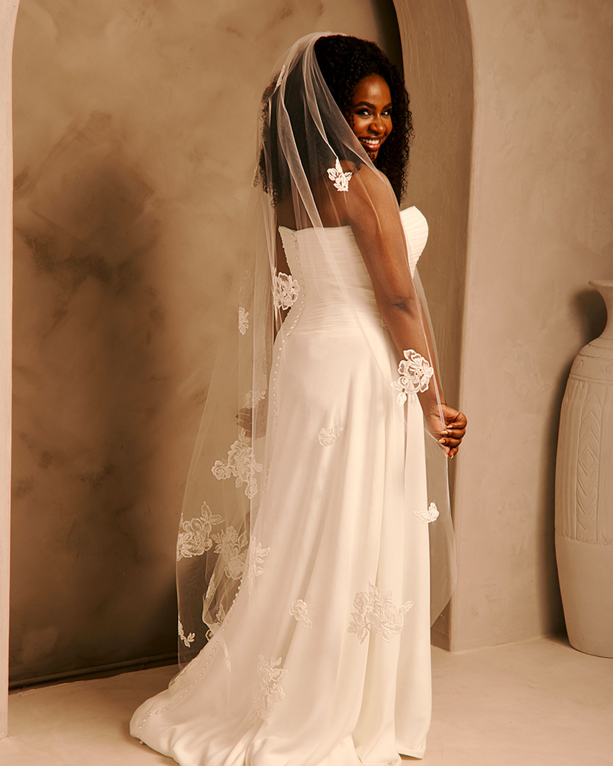 Bridal Veil with Painted Flowers and Pearls Cassandra Lynne - A woman wearing a strapless ivory wedding gown with floral lace appliques and a long sheer veil, photographed in a warm beige studio setting.