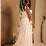 Bridal Veil with Painted Flowers and Pearls Cassandra Lynne - A woman wearing a strapless ivory wedding gown with floral lace appliques and a long sheer veil, photographed in a warm beige studio setting.
