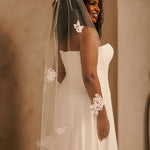 Bridal Veil with Painted Flowers and Pearls Cassandra Lynne - A woman wearing a white strapless wedding gown and a sheer ivory veil with floral lace appliques, photographed indoors against a warm beige wall.
