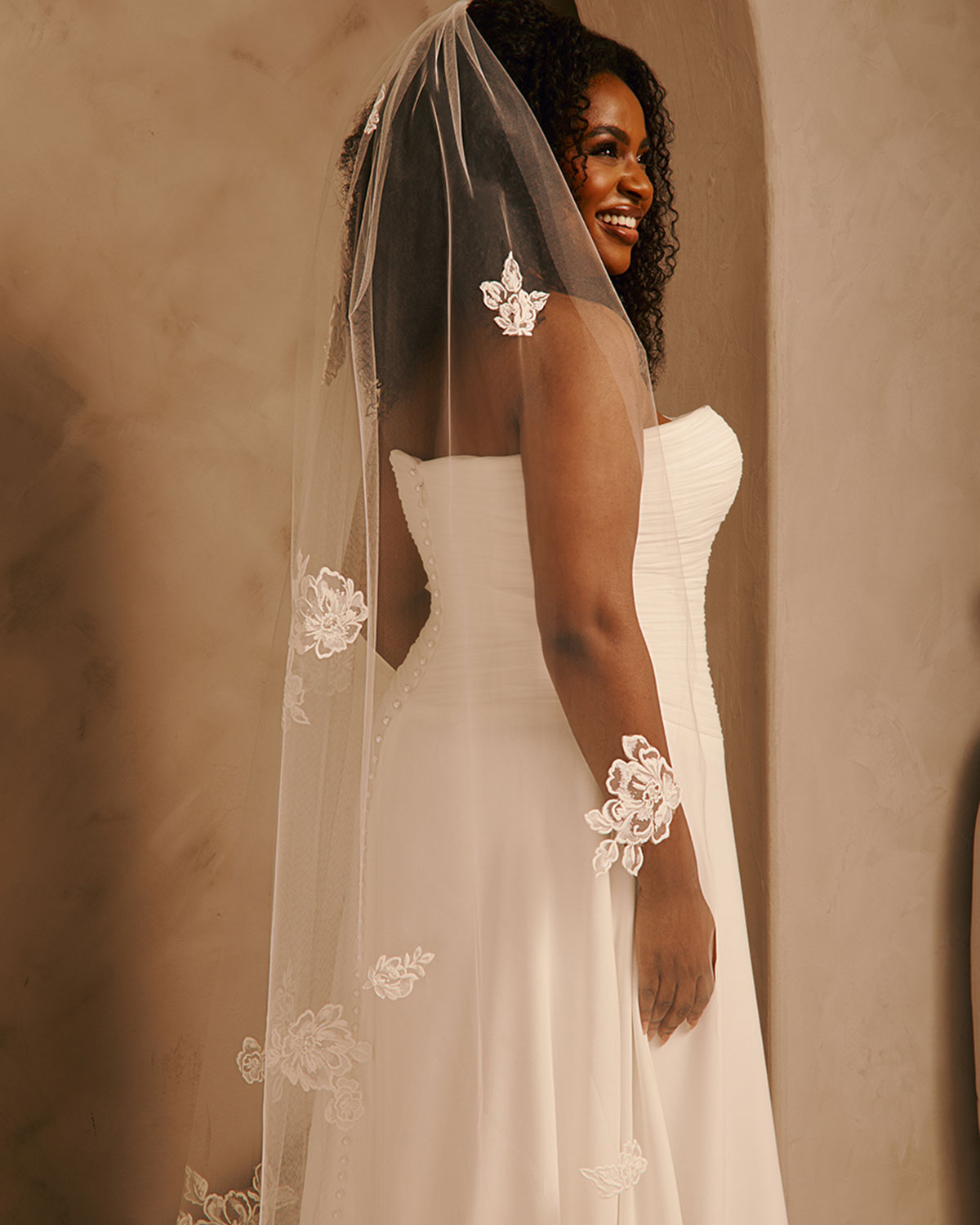 Bridal Veil with Painted Flowers and Pearls Cassandra Lynne - A woman wearing a white strapless wedding gown and a sheer ivory veil with floral lace appliques, photographed indoors against a warm beige wall.