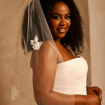 Bridal Veil with Painted Flowers and Pearls Cassandra Lynne - A smiling woman with dark curly hair wears a sheer ivory bridal veil with silver floral embroidery and a white strapless wedding dress, posed against a warm beige stone wall.